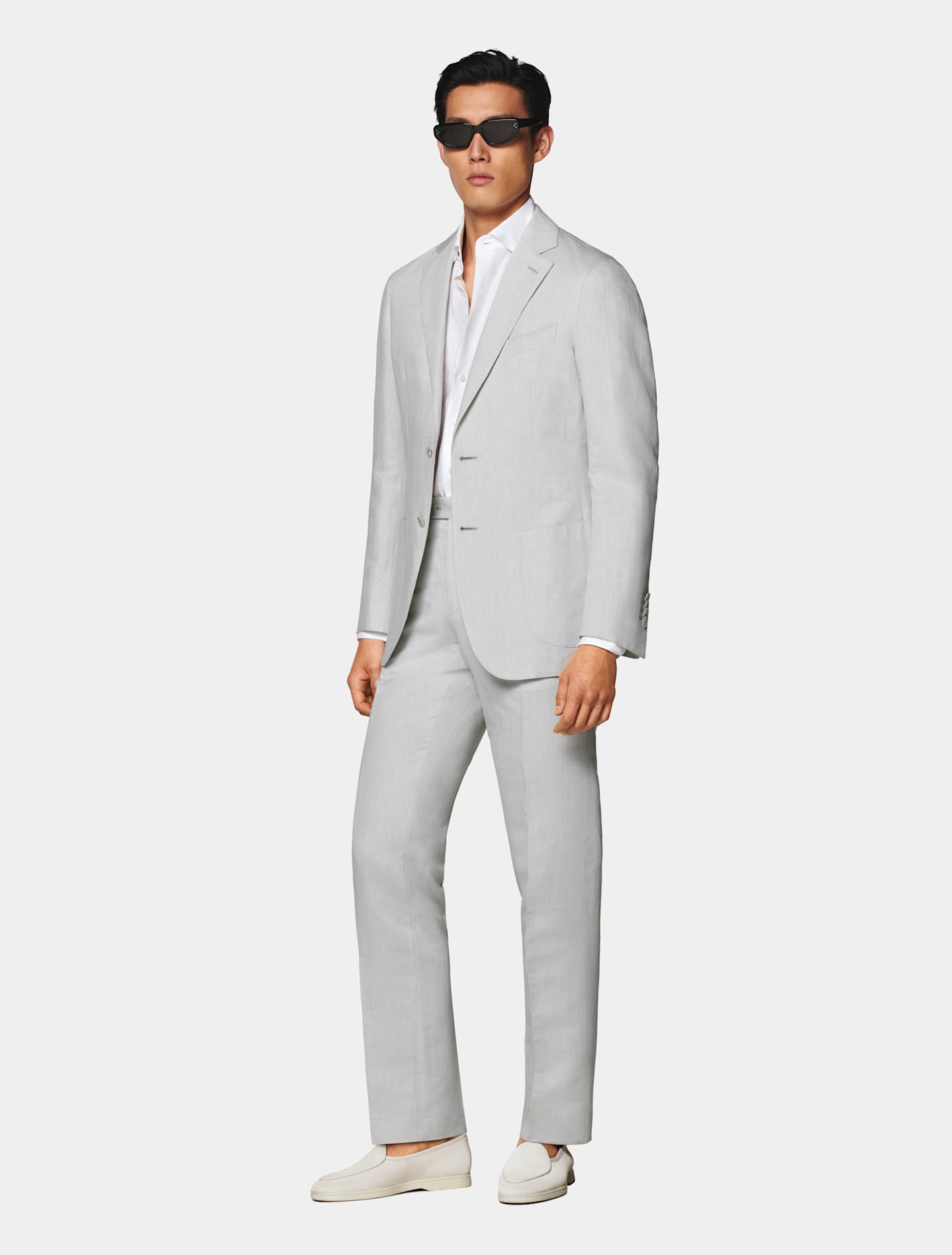 Enkelknäppt ljusgrå kostym buren med löst knäppt vit skjorta och ljust mullvadsfärgade loafers.