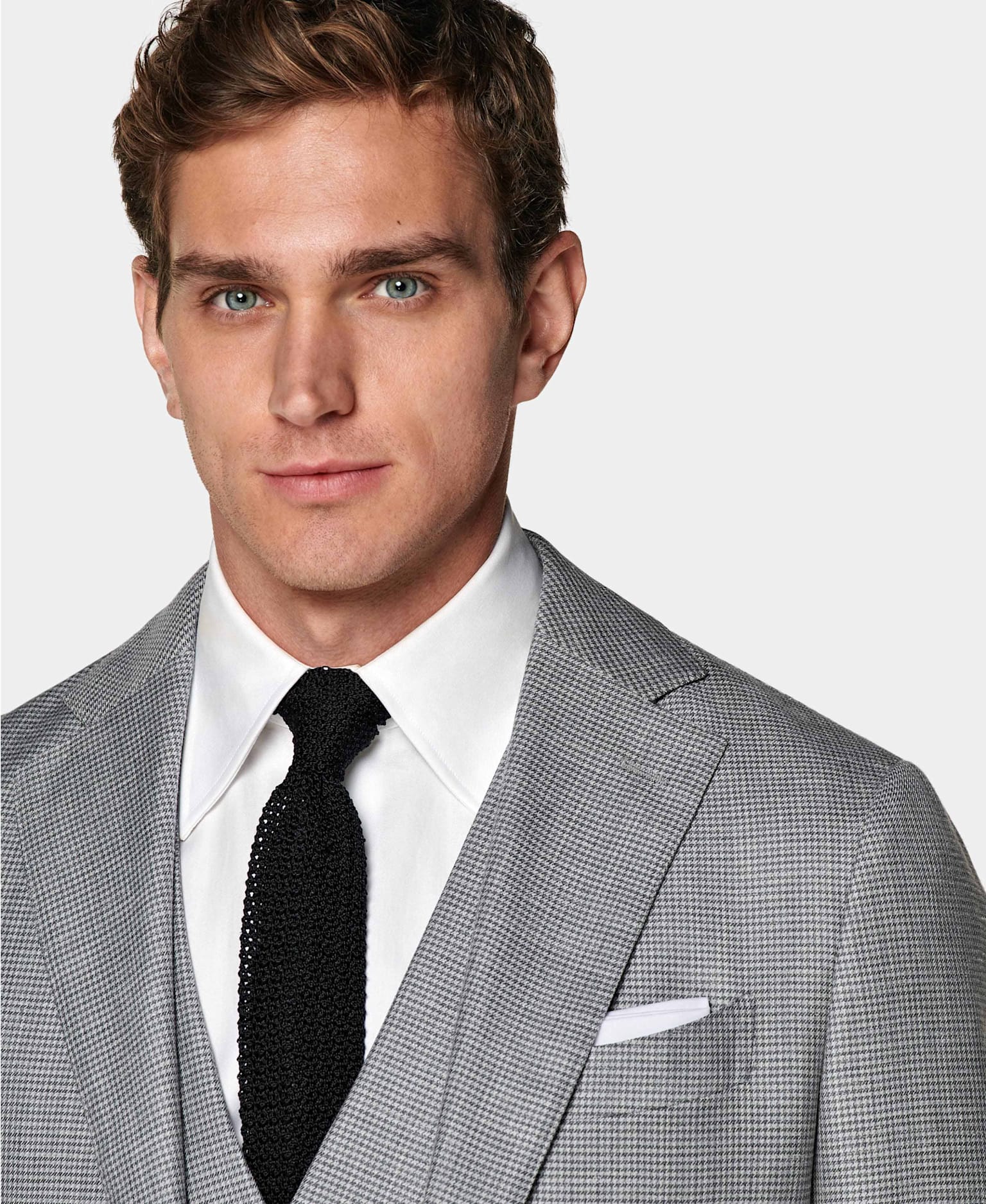 Dettaglio di abito tre pezzi grigio con quadretto pied-de-poule, camicia bianca, cravatta nera in seta lavorata a maglia, e pochette bianca.